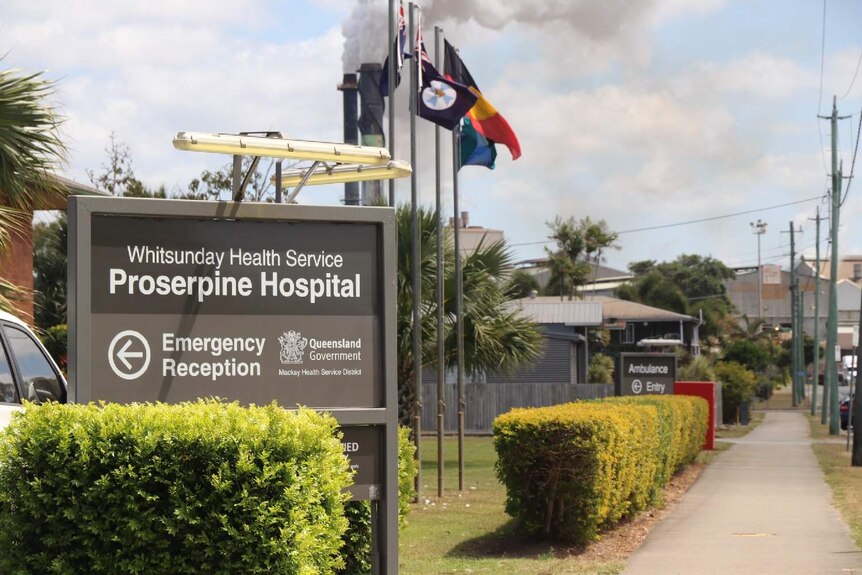 Sign for the Proserpine Hospital outside the building with flags on the property and sugar mill smoke stacks in the distance.