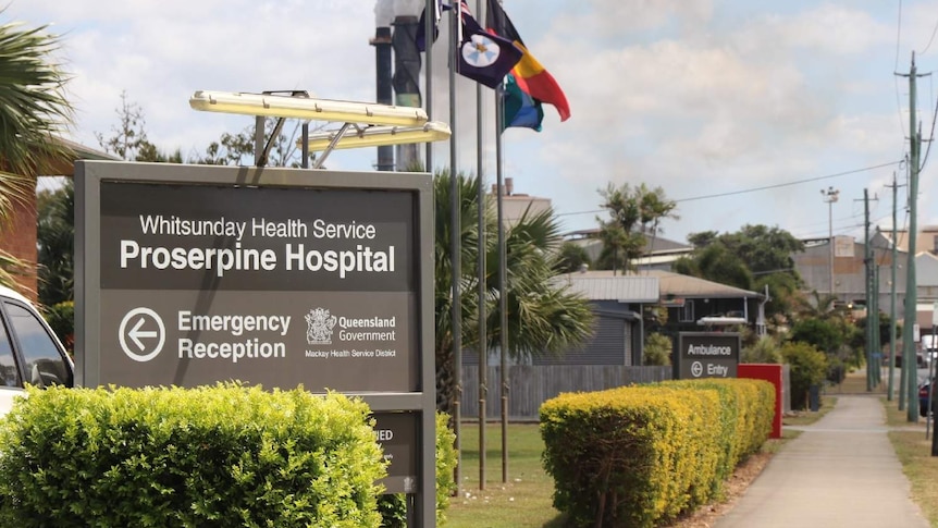 Sign for the Proserpine Hospital outside the building with flags on the property and sugar mill smoke stacks in the distance.