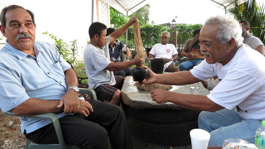 Peter Christian sits as a table in an outdoor setting and is passed a cup of kava.
