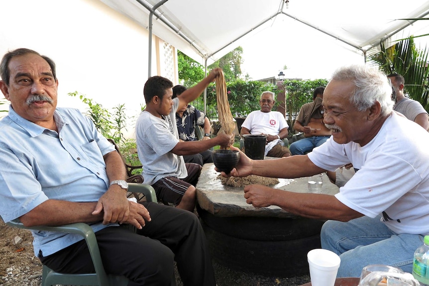 Peter Christian sits as a table in an outdoor setting and is passed a cup of kava.
