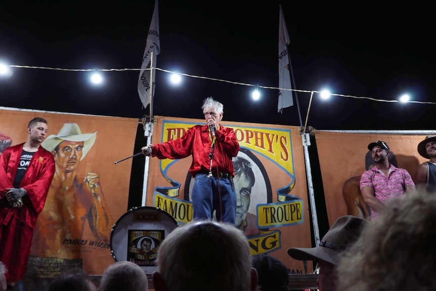 An older man in a red silk shirt and jeans stands on a stage with a string of bulbs and a colorful banner, speaks into a microphone.