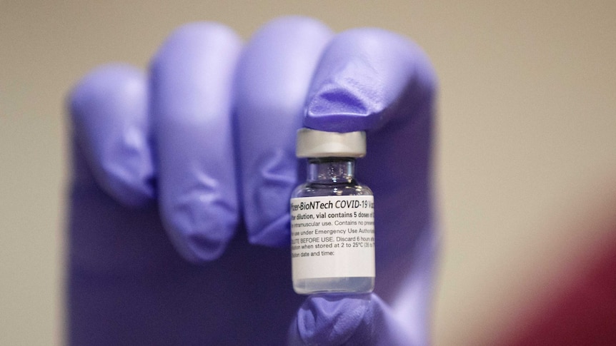 A gloved hand holds up a small vial of Pfizer's COVID-19 vaccine