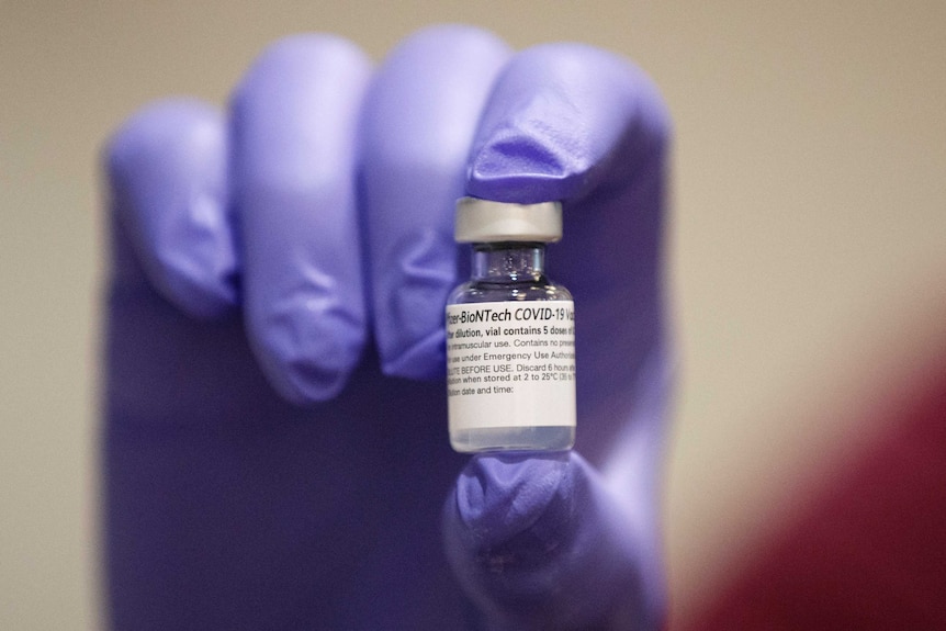 A gloved hand holds a small vial of Pfizer's COVID-19 vaccine