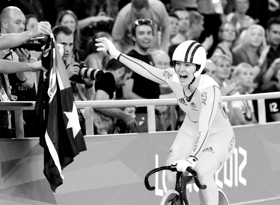 Anna Meares on her bike smiling and celebrating a win with an Australian flag in the foreground.