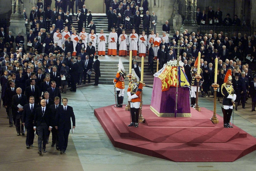 Former British prime minister Tony Blair walks past the Queen Mother's coffin as she lies in state under a tent, guarded by men.