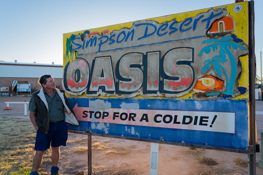 A man stands next to and looks at a colourful, peeling sign that says Simpson Desert Oasis - Stop for a coldie!