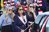 Four women holding walk behind a hearse draped with St Kilda football club flags.