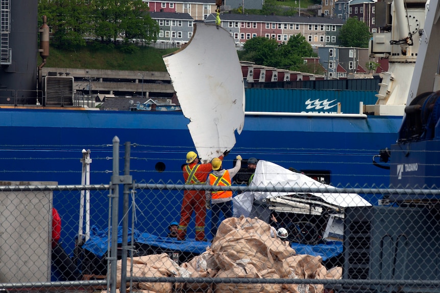 A blue ship is pictured as a large white piece of debris is being unloaded by men wearing high-vis and helmets.