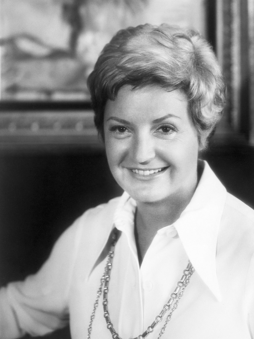 A woman with short cropped hairstyle, wearing chains over a crisp white shirt, smiles in black and white portrait