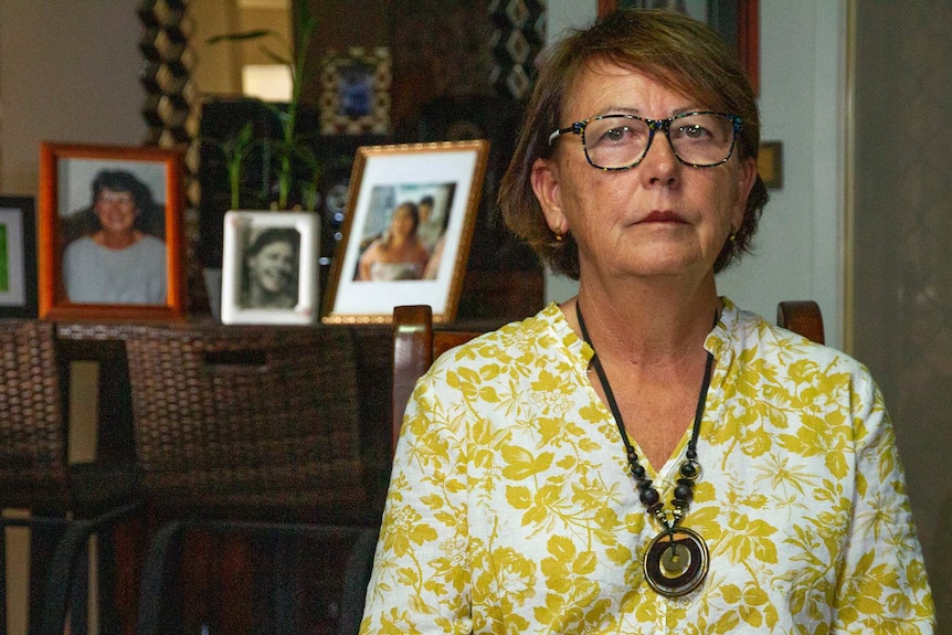 A woman wearing a white and yellow shirt sits in front of family photos.