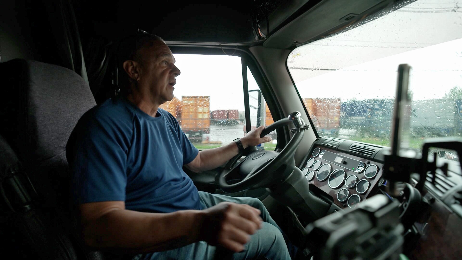 A man sits in the left-side driver's seat of a large truck. Outside shipping containers can be seen.
