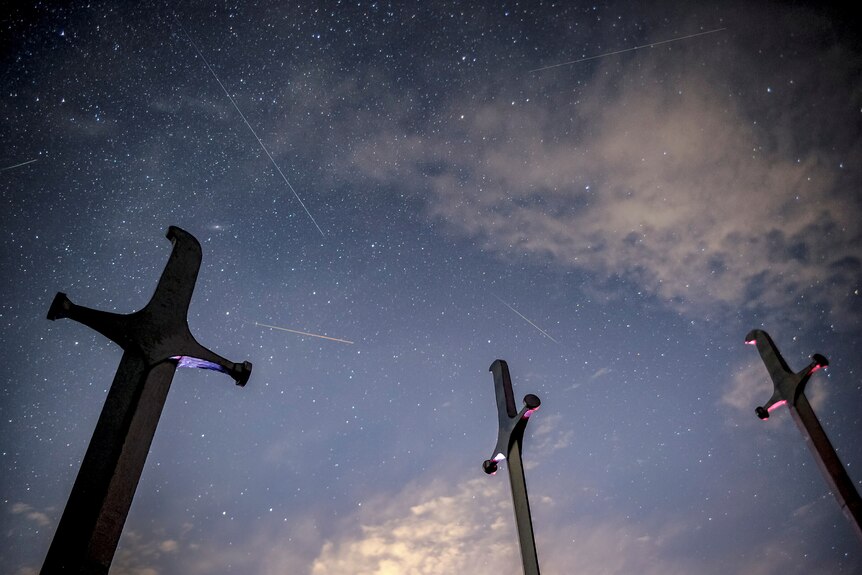 Meteors streak past stars in the night sky behind stone crosses.
