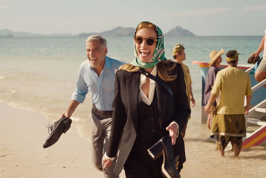 Middle-aged white man with grey hair wears blue shirt behind woman with red hair, sunglasses, head scarf and suit on a beach.