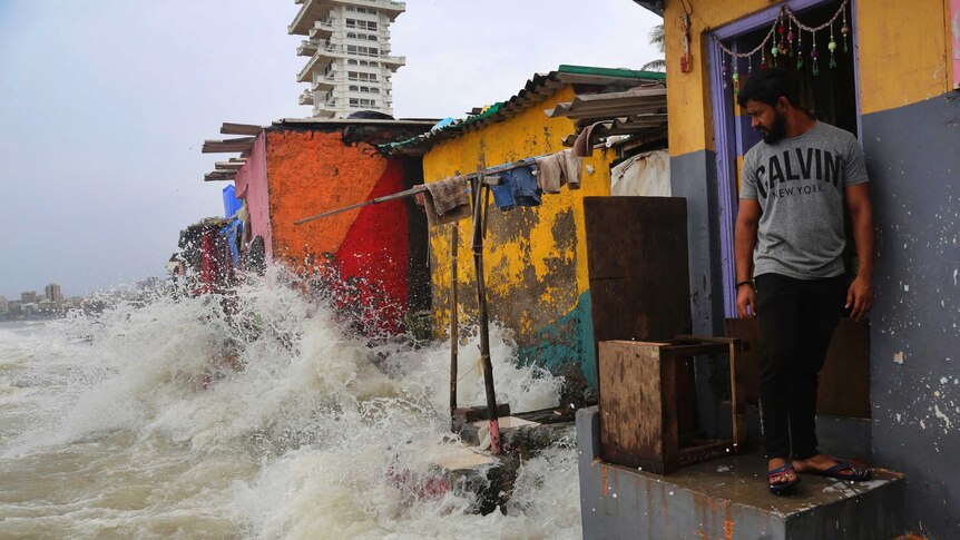 A man stands on a stoop and looks down as the sea crashes into brightly painted huts on Mumbai's coastline.