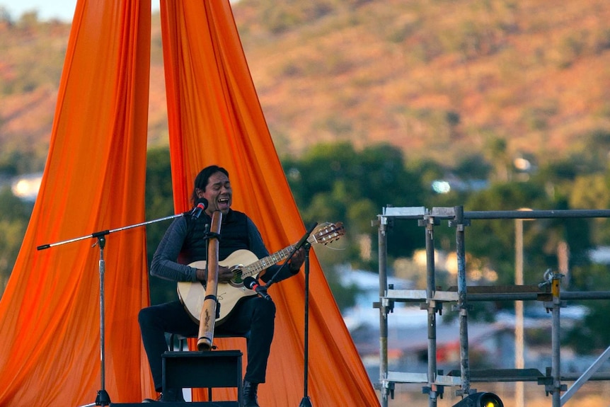 A indigenous man sits on a stage playing the guitar and singing, with a didgeridoo in front of him.