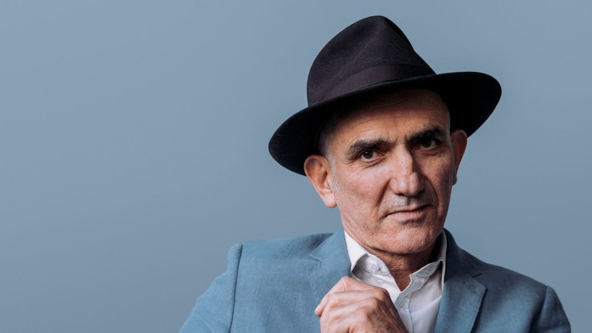 A portrait of Australian musician Paul Kelly who is wearing a pale blue-grey suit and black hat.