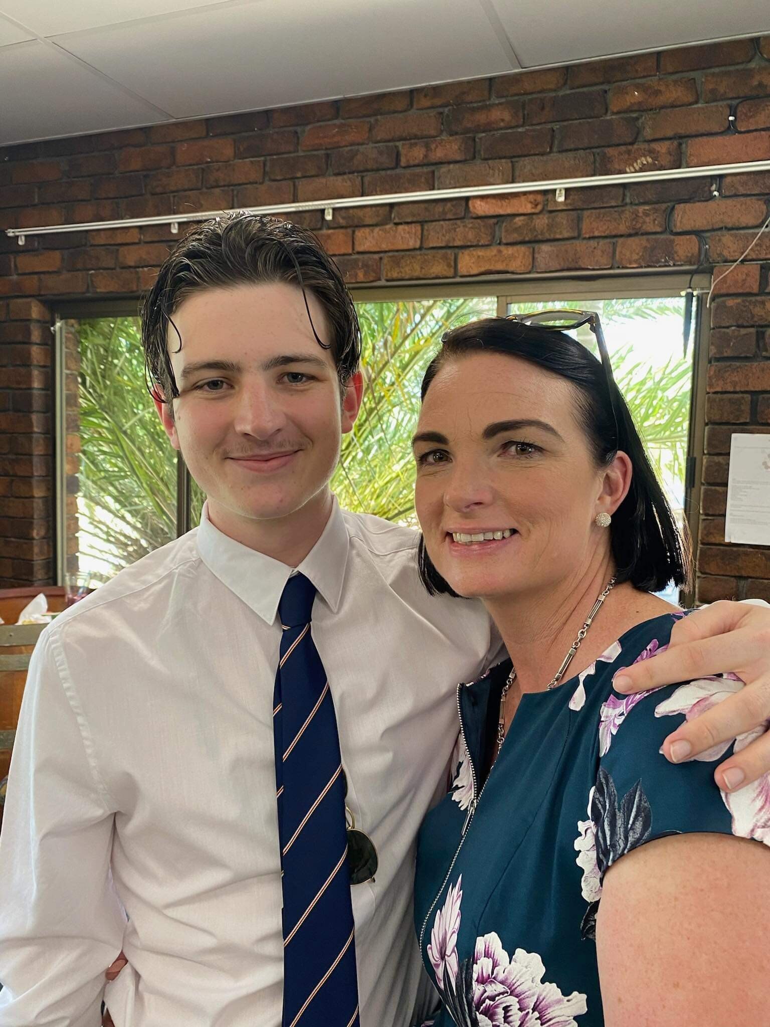 A teenage boy wearing a white shirt and navy tie with his arm around his mum with short dark hair.