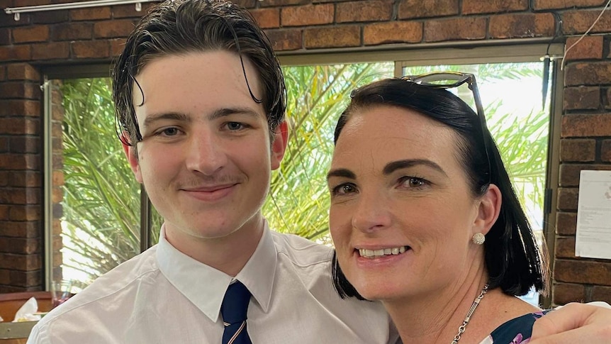 A teenage boy wearing a white shirt and navy tie with his arm around his mum with short dark hair.