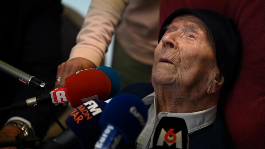 A very elderly person leans their head back on a red pillow and microphones from various media are held in front of her.