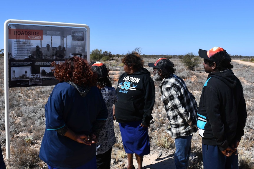 A group of five people stand reading a sign in the outback.