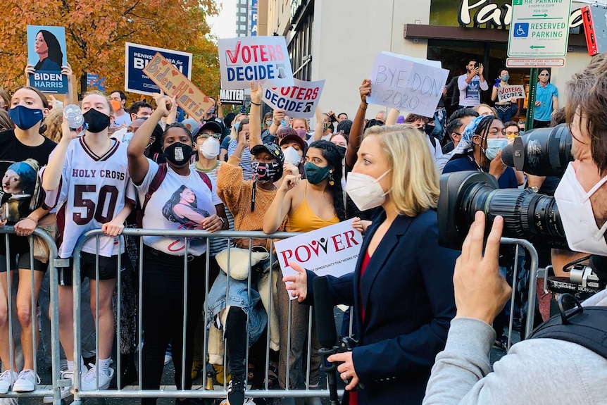 Cameraman filming woman wearing a mask in front of crowd of people.