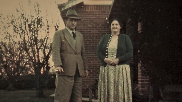 A man and woman stand outside a red brick house in the 60s.