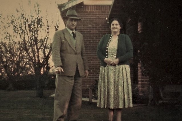 A man and woman stand outside a red brick house in the 60s.