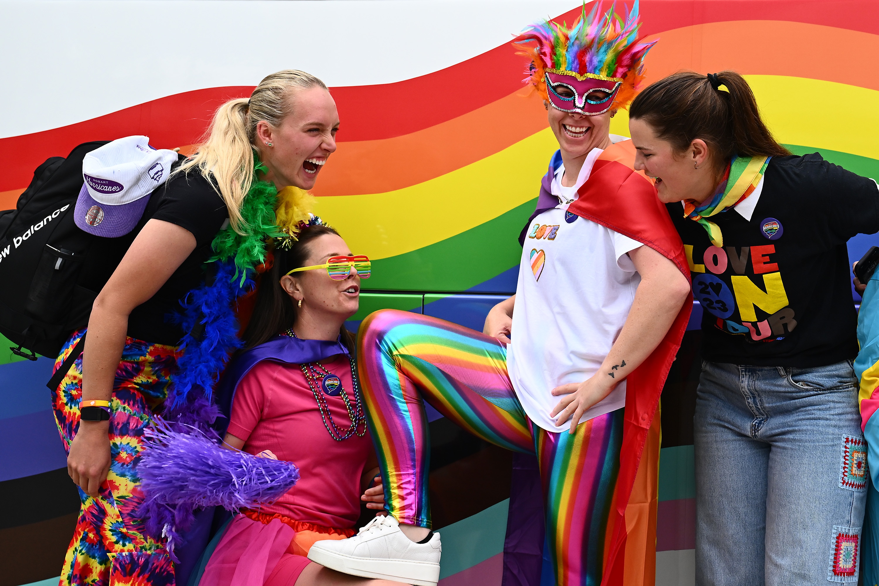 Four WBBL players stand in front of a rainbow mural, they're all wearing bright, rainbow clothes and smiling for camera