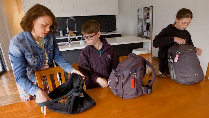 A woman and two children in school uniforms pack bags at a table.