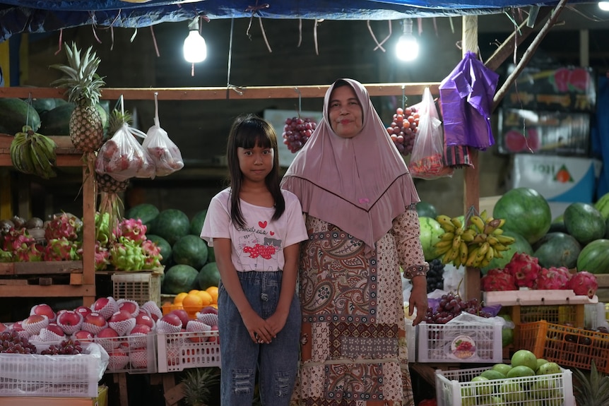 Madre con hijab e hija paradas frente a un puesto de frutas sonriendo mirando a la cámara.
