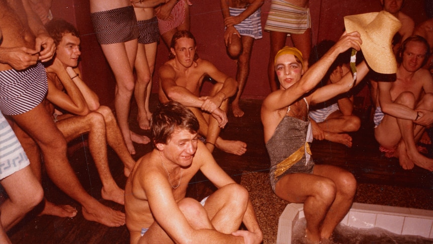Men in bathers sit around a sauna together laughing