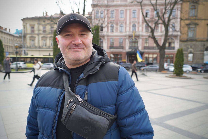 A man wearing a dark blue puffer jacket and cap smiles while wearing a bumbag.