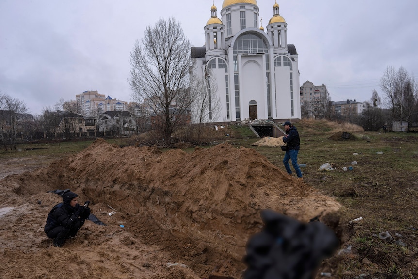 Journalists take pictures next to a mass grave and a church