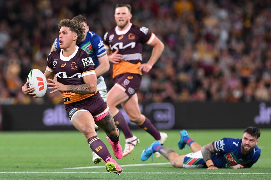 An NRL player running with the ball, one defender on the ground, and another chasing.