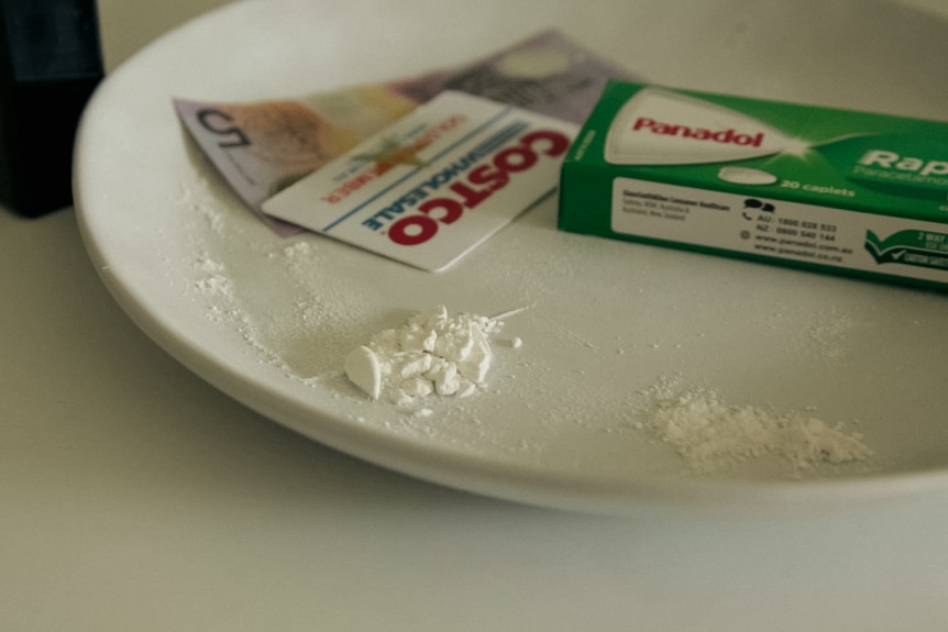 Clumps of white powder on a dinner plate. Behind it sit a box of panadol, a Costco plastic card, and a $5 note.
