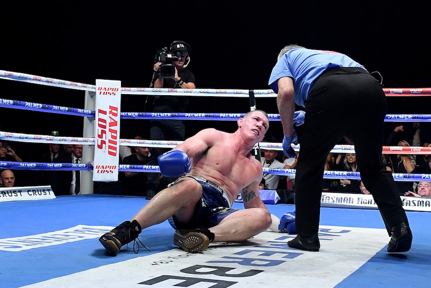 Paul gallen looks up at a referee as he lies in a boxing ring, who is bending over him and pointing at him
