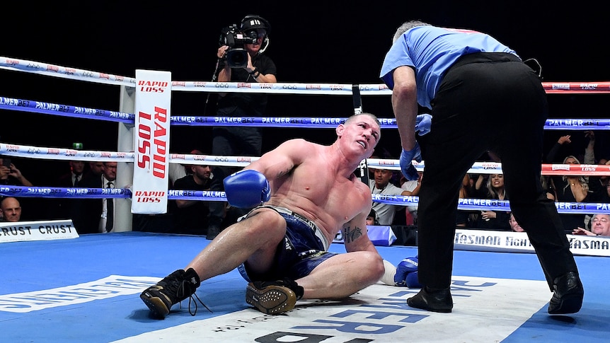 Paul gallen looks up at a referee as he lies in a boxing ring, who is bending over him and pointing at him