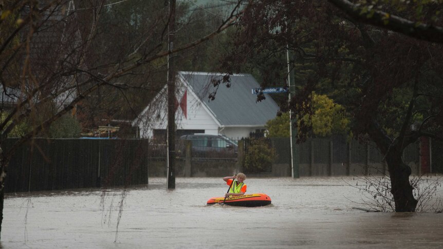 Person paddles a brightly coloured inflatable canoe in a Christchurch street flooded with murky water