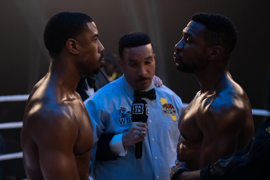 Two shirtless men in a boxing ring stare at each other, while a man between them holds a microphone.