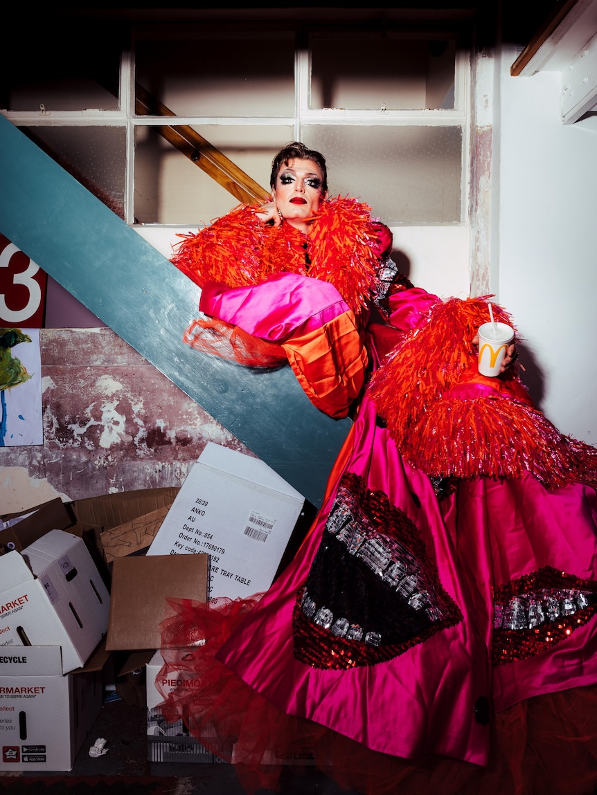 A man in his 30s with a full face of drag makeup, in a larfe pink and orange feather cape, holding a mcdonalds drink on a stair