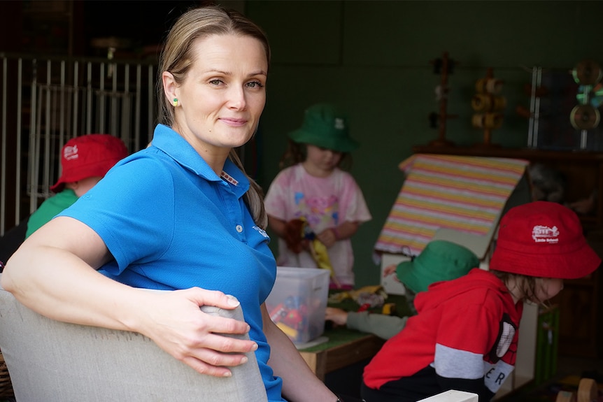 a woman sits on a chair and smiles at the camera as children play behind her