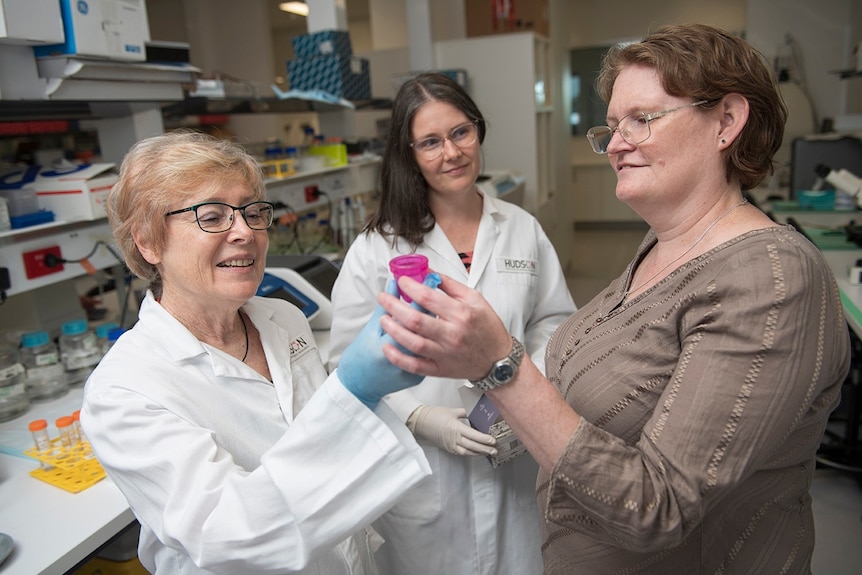 Профессор Гаргетт и Николь Фернли держат розовую менструальную чашу в лаборатории под наблюдением коллеги.