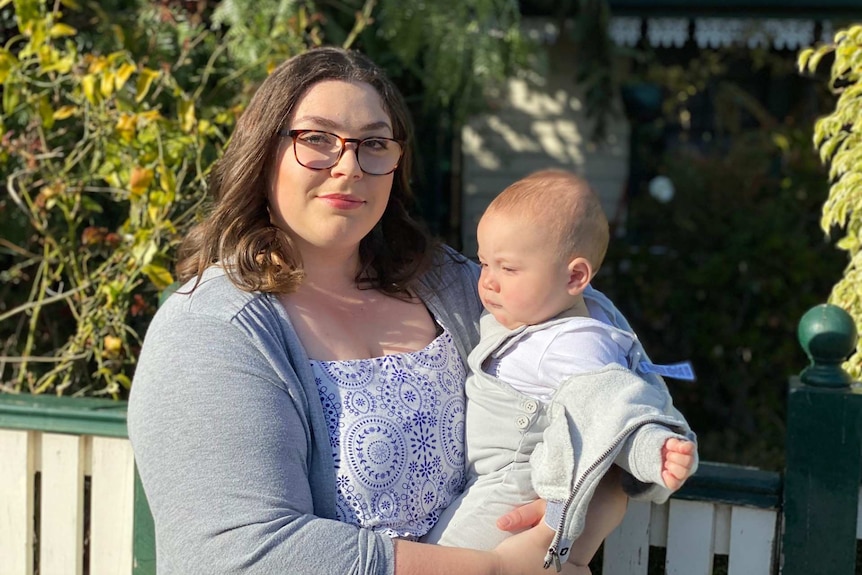 A woman wearing glasses looks into the camera as she holds a baby boy while standing at her front fence.