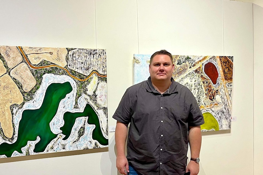A First Nations man stands in front of paintings in an exhibition