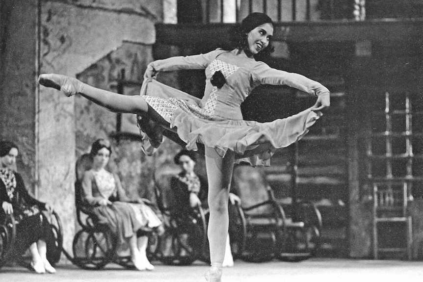 A ballet dancer on stage, she stands on one leg and holds her skirt out