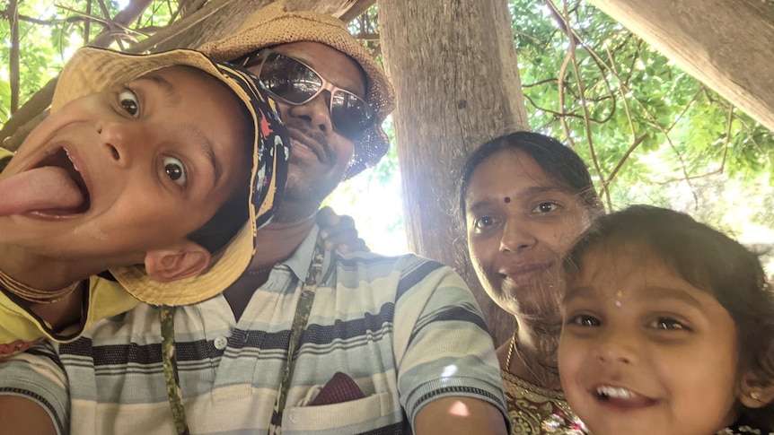 Vinu, Akila and their two children taking a selfie, smiling and pulling faces, shot from below with green foliage behind them