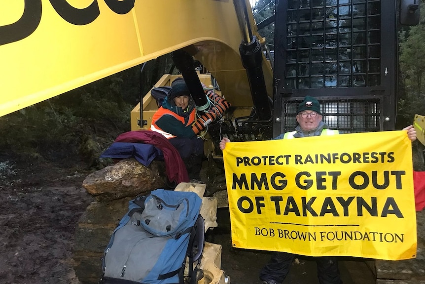 Two people sit on machinery, one holding a sign saying MMG Get out of Takayna.