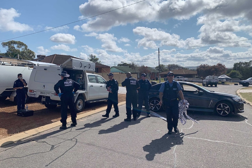 Police forensic officers stand on a road with another officer walking towards the camera away from a blue car.