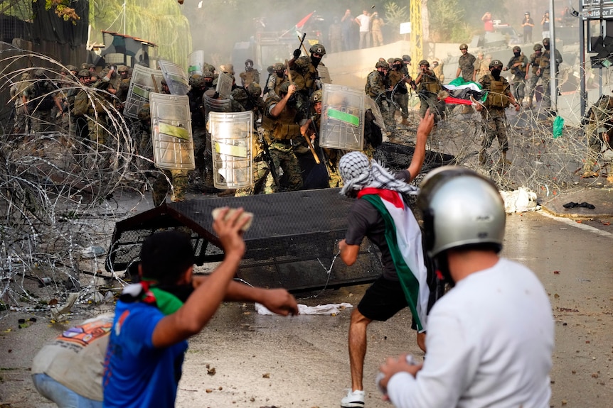 Three young men throw stones towards soldiers using riot shields to form a blockade during a demonstration.