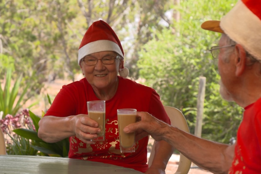 Elderly couple Marg and Ed, in red santa hats and red christmas shirts, cheers a glass of iced coffee in the shady patio.